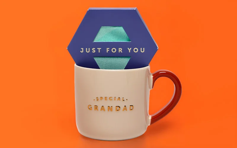 A cream ceramic mug with a brown handle, printed with the text ‘Special Grandad’ in gold lettering, holding a navy gift box labelled ‘Just for You’ with a pair of green socks visible through cut‑out windows, displayed against a bright orange background. A cream ceramic mug with a brown handle, printed with the text ‘Special Grandad’ in gold lettering, holding a navy gift box labelled ‘Just for You’ with a pair of green socks visible through cut‑out windows, displayed against a bright orange background.