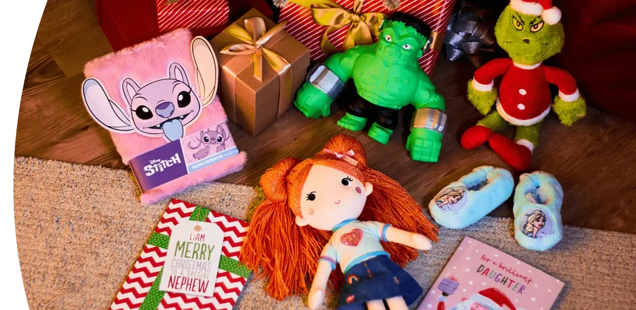 Christmas gifts arranged on a wooden floor, including a pink Stitch notebook, wrapped presents, a green Hulk figure, a Grinch plush toy in a Santa outfit, a doll with orange yarn hair, blue Frozen slippers, and two festive greeting cards.