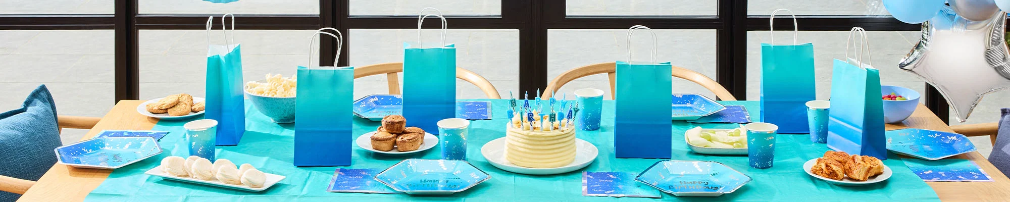 A blue-themed children's birthday party table laid with ombré blue table covers, matching party bags, paper cups, plates and napkins. A birthday cake topped with blue candles sits at the centre, surrounded by party food including cookies, meringues, pastries and fruit. The table is arranged in front of large windows and decorated for a coordinated blue birthday celebration. A blue-themed children's birthday party table laid with ombré blue table covers, matching party bags, paper cups, plates and napkins. A birthday cake topped with blue candles sits at the centre, surrounded by party food including cookies, meringues, pastries and fruit. The table is arranged in front of large windows and decorated for a coordinated blue birthday celebration.