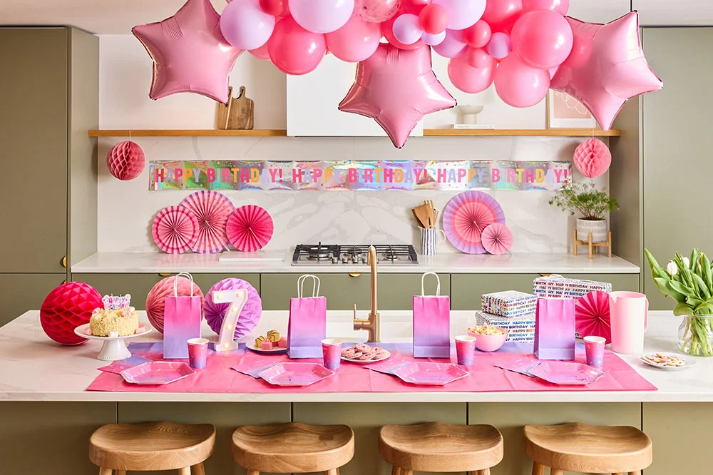 A pink-themed birthday party setup in a modern kitchen featuring a large balloon garland with pink star-shaped foil balloons, a holographic ‘Happy Birthday’ banner and pink paper fan decorations. The table is laid with ombré pink and purple partyware, gift bags, wrapped presents, sweet treats, a birthday cake and illuminated number 7 lights, creating a coordinated seventh birthday celebration.