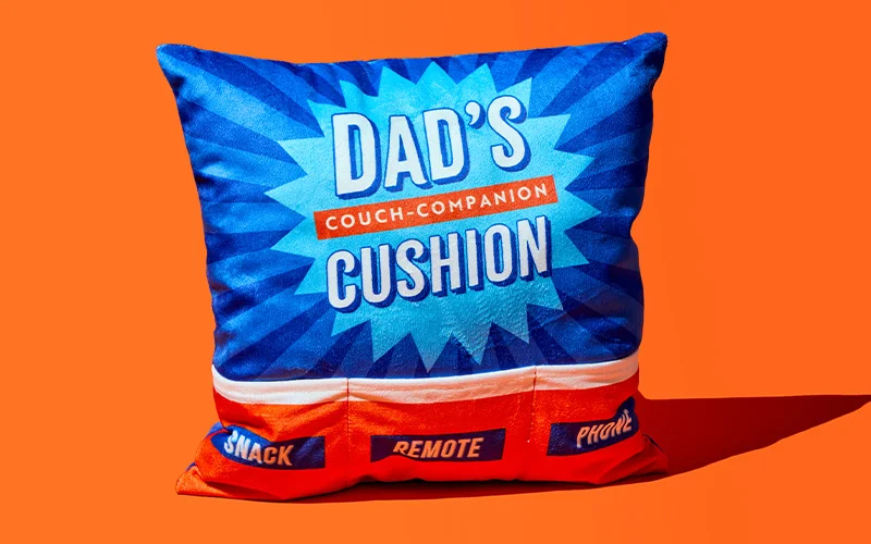 A square novelty cushion in blue and red with a starburst design and bold text reading ‘Dad’s Couch‑Companion Cushion,’ featuring built‑in compartments labelled ‘Snack,’ ‘Remote,’ and ‘Phone,’ displayed against a bright orange background. A square novelty cushion in blue and red with a starburst design and bold text reading ‘Dad’s Couch‑Companion Cushion,’ featuring built‑in compartments labelled ‘Snack,’ ‘Remote,’ and ‘Phone,’ displayed against a bright orange background.
