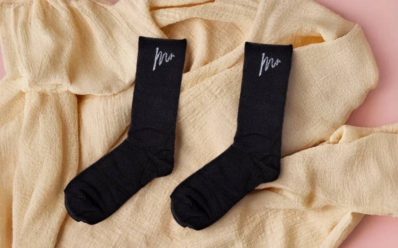 A pair of black socks with ‘Mr’ printed in white script near the top, laid flat on soft beige fabric. A pair of black socks with ‘Mr’ printed in white script near the top, laid flat on soft beige fabric.