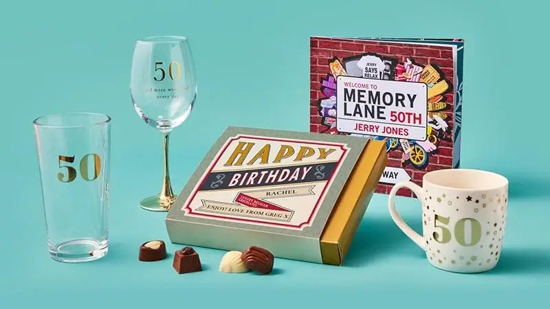 A collection of 50th birthday-themed items on a teal background, including a wine glass, a mug, a book, a gift box labeled 'Happy Birthday Rachel', and chocolates