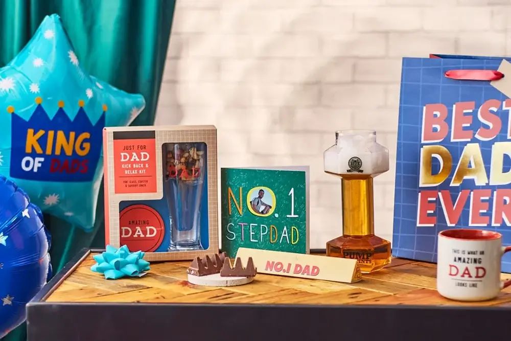 The image shows a Father's Day gift setup, including two greeting cards, a mug with 'Best Dad Ever' written on it, a wooden crate labeled 'Dad's Snack Crate' filled with snacks and drinks, and a small bowl of mixed nuts. The image shows a Father's Day gift setup, including two greeting cards, a mug with 'Best Dad Ever' written on it, a wooden crate labeled 'Dad's Snack Crate' filled with snacks and drinks, and a small bowl of mixed nuts.