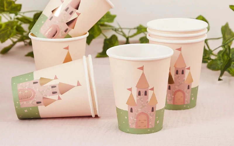 A set of pastel pink paper cups featuring an illustrated fairytale castle with glitter-effect accents and green decorative details, displayed on a table with trailing greenery in the background. A set of pastel pink paper cups featuring an illustrated fairytale castle with glitter-effect accents and green decorative details, displayed on a table with trailing greenery in the background.