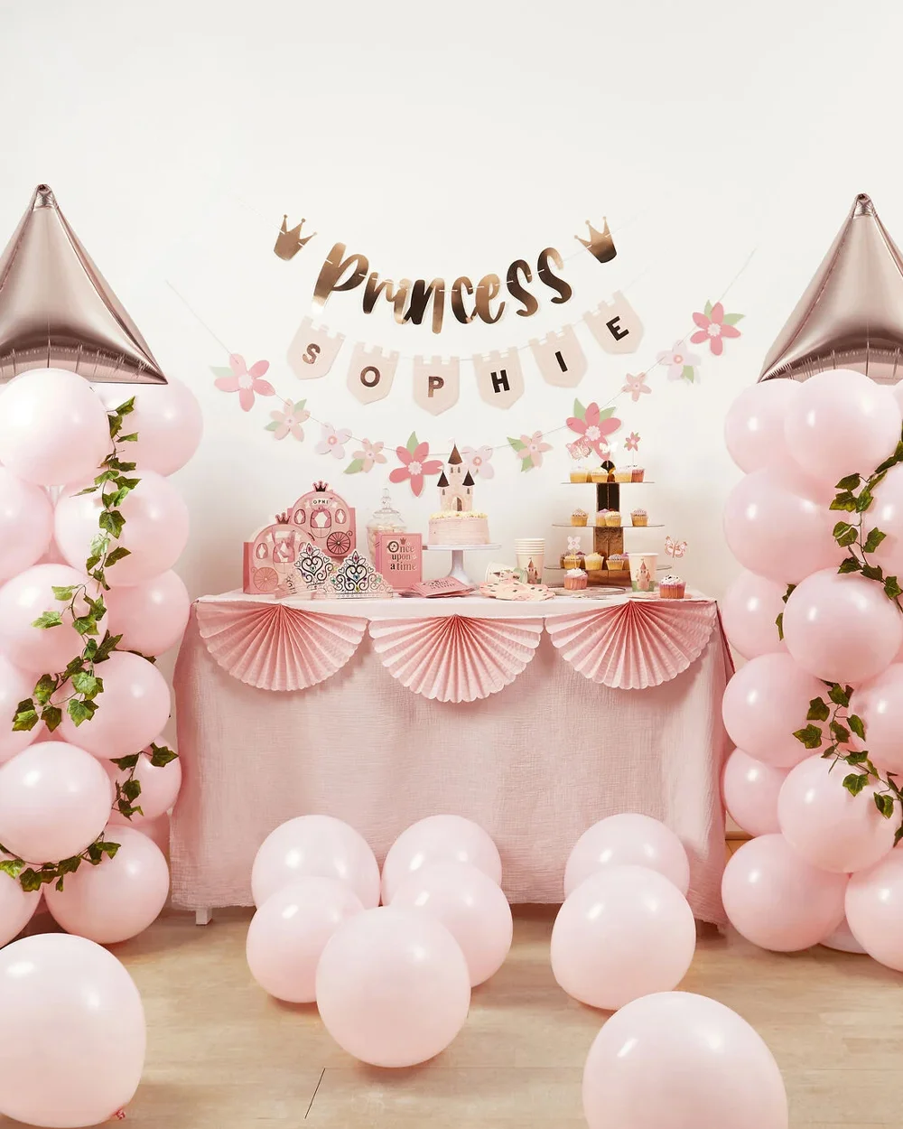 A princess-themed party table decorated in pastel pink, featuring balloon columns topped with rose-gold castle-turret foil balloons and entwined with green foliage. Above the table hangs a personalised banner reading ‘Princess Sophie,’ alongside floral garlands and crown decorations. The table is styled with a celebration cake, cupcakes, party tableware, favour boxes and fairytale-themed accessories, with pale pink balloons scattered across the floor in front.