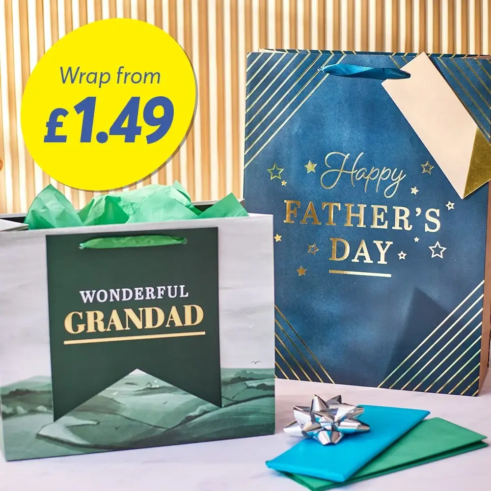The image shows two gift bags, one green and white with the text 'WONDERFUL GRANDAD' and the other blue with gold accents and the text 'Happy FATHER'S DAY.' There is also a yellow circle with the text 'Wrap from £1.49' in blue. A small silver bow and two pieces of colored paper are placed in front of the bags.