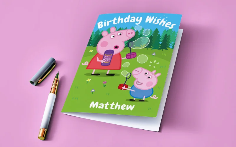 A personalised Peppa Pig birthday card titled ‘Birthday Wishes,’ featuring Peppa Pig and a smaller character blowing bubbles in a green outdoor scene, with the name ‘Matthew’ printed at the bottom. A pen is placed beside the card on a pink background.