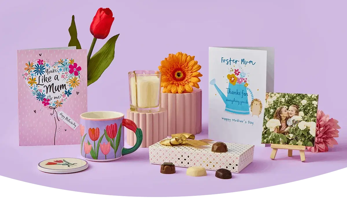 A cheerful Mother’s Day display featuring two greeting cards, a red tulip, a floral mug with a matching coaster, a candle, a gift box with assorted chocolates, and a small photo plaque arranged on a lilac background. A cheerful Mother’s Day display featuring two greeting cards, a red tulip, a floral mug with a matching coaster, a candle, a gift box with assorted chocolates, and a small photo plaque arranged on a lilac background.