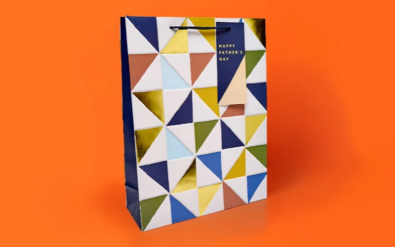 A large Father’s Day gift bag with a geometric triangle pattern in blue, gold, white and green, featuring a small tag reading ‘Happy Father’s Day,’ displayed against a bright orange background. A large Father’s Day gift bag with a geometric triangle pattern in blue, gold, white and green, featuring a small tag reading ‘Happy Father’s Day,’ displayed against a bright orange background.
