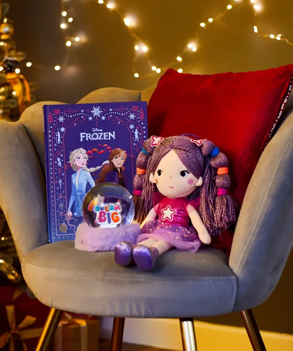 Cozy chair with a red cushion displaying a Disney Frozen book with Elsa and Anna, a plush doll with purple hair and a star on its dress, and a snow globe with Dream Big on a fluffy base, set against a background of warm fairy lights.