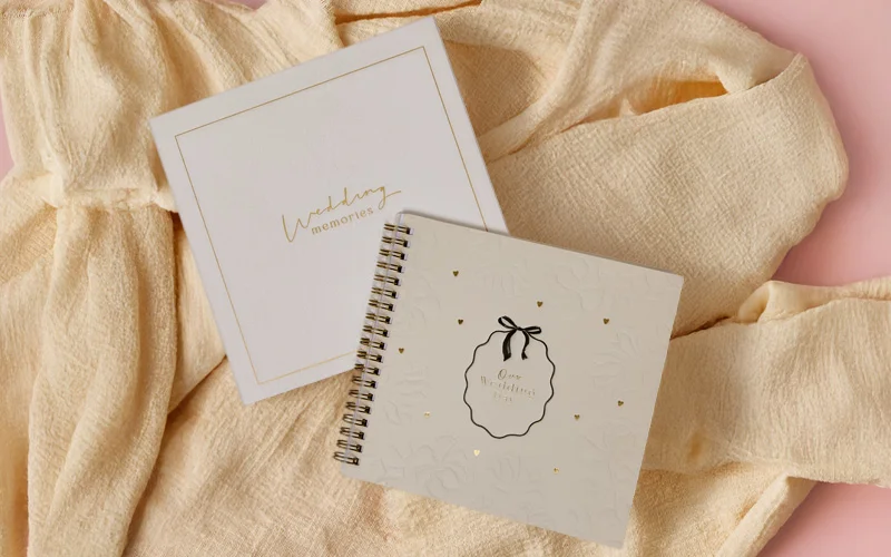 “A set of wedding keepsake books on beige fabric, including a ‘Wedding memories’ box and a spiral‑bound album with gold foil hearts and a ribbon illustration, displayed on a pink background. “A set of wedding keepsake books on beige fabric, including a ‘Wedding memories’ box and a spiral‑bound album with gold foil hearts and a ribbon illustration, displayed on a pink background.