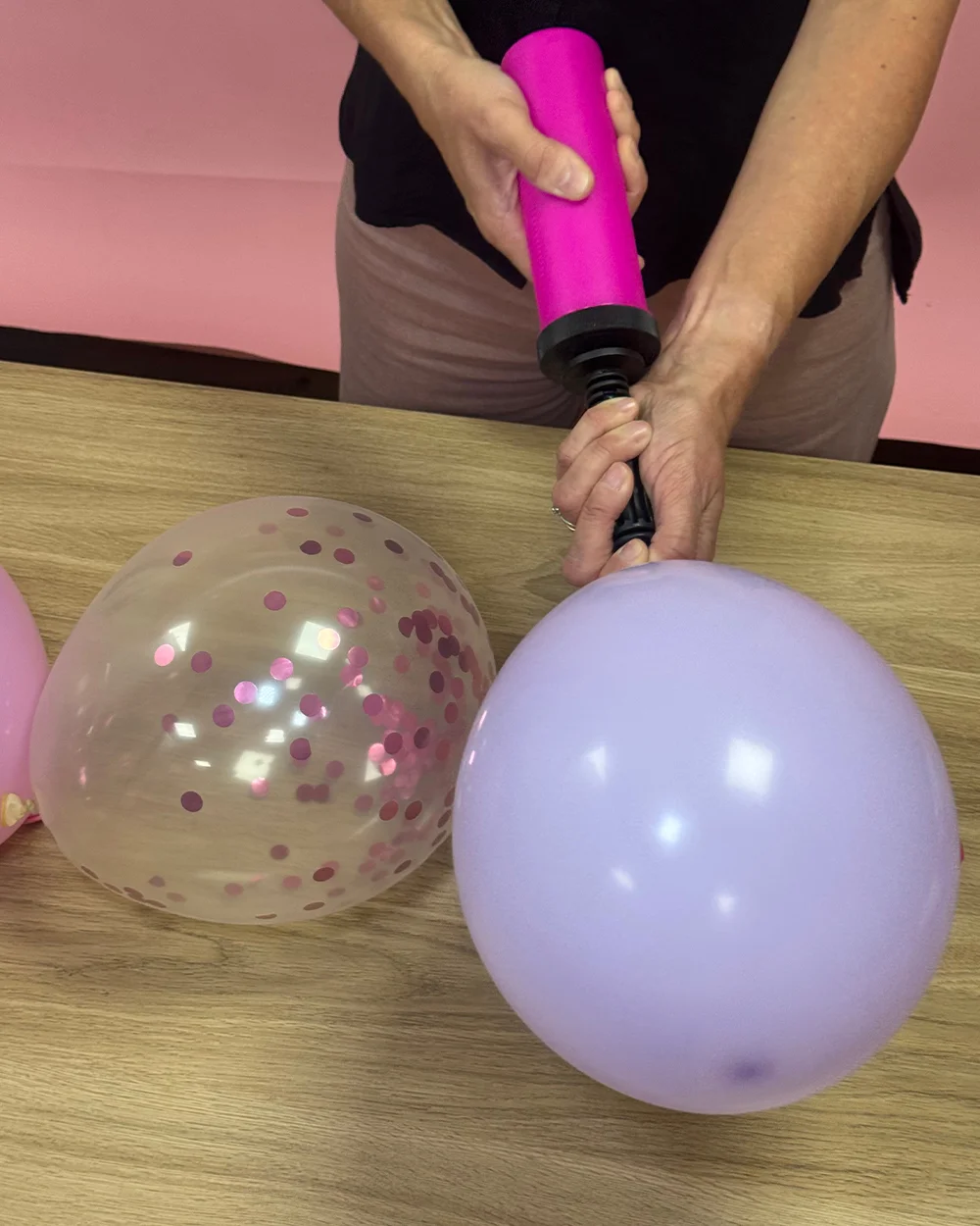 A balloon arch tutorial step showing a pink hand pump being used to inflate a lilac balloon on a wooden tabletop. A clear balloon filled with pink confetti and additional pink balloons are positioned nearby, demonstrating how to prepare and inflate the different balloons needed to create a balloon arch display. A balloon arch tutorial step showing a pink hand pump being used to inflate a lilac balloon on a wooden tabletop. A clear balloon filled with pink confetti and additional pink balloons are positioned nearby, demonstrating how to prepare and inflate the different balloons needed to create a balloon arch display.