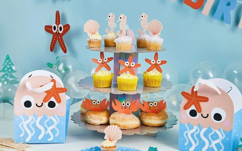 An under-the-sea themed party display featuring a three-tier stand filled with cupcakes and doughnuts decorated with seahorse, shell, starfish and crab toppers. Jellyfish-shaped party favour boxes, clear balloons and ocean-themed decorations complete the setup against a light blue background.