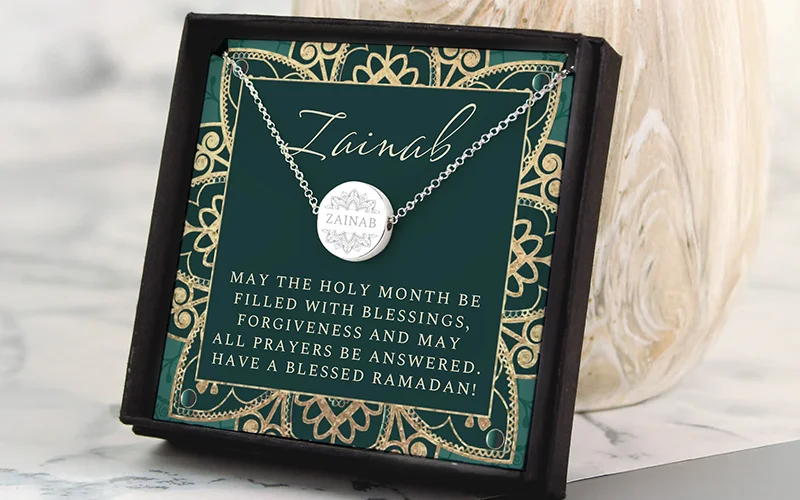 A personalised silver necklace displayed in a dark gift box, featuring a round pendant engraved with the name ‘Zainab’ and a Ramadan message on a green and gold decorative background.