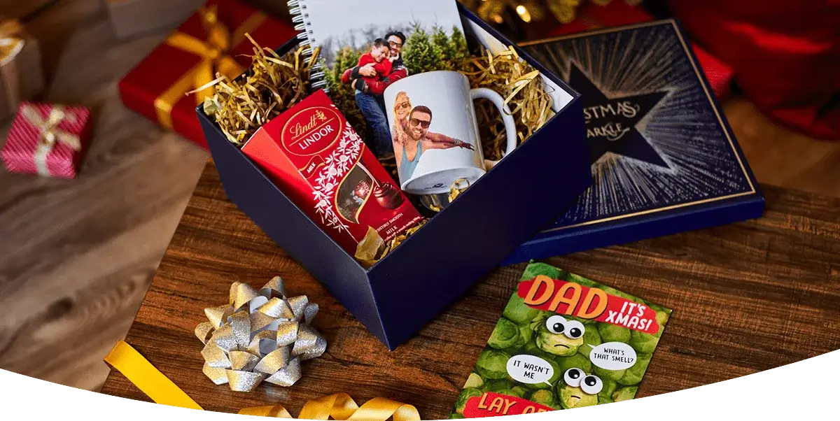 Festive holiday display with greeting cards, themed mugs labeled Mummy Claus, Daddy Claus, and Mini Claus, candles, pen, gold ribbon, and Christmas ornaments against a red background. Festive holiday display with greeting cards, themed mugs labeled Mummy Claus, Daddy Claus, and Mini Claus, candles, pen, gold ribbon, and Christmas ornaments against a red background.