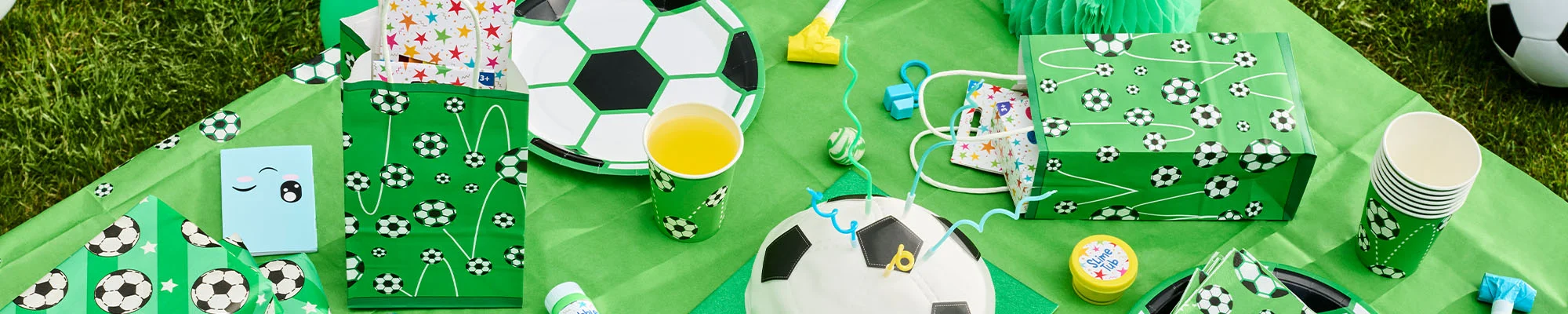 A football-themed party table display arranged on a green tablecloth, featuring football-print gift bags, matching paper plates, cups and napkins, party favour bags filled with small gifts, a football-shaped cake, and party bag fillers including bubbles, slime and novelty toys. The setup is styled outdoors on grass with football-themed decorations throughout. A football-themed party table display arranged on a green tablecloth, featuring football-print gift bags, matching paper plates, cups and napkins, party favour bags filled with small gifts, a football-shaped cake, and party bag fillers including bubbles, slime and novelty toys. The setup is styled outdoors on grass with football-themed decorations throughout.