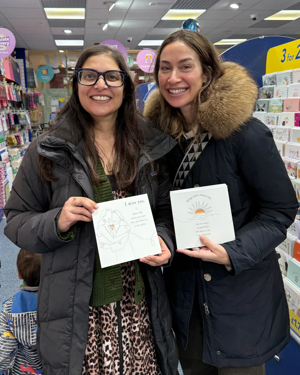 Two people inside a greeting‑card shop, each holding a Mother’s Day card. One card displays a line‑art illustration with a heart cut‑out and the message ‘I miss you.’ The other shows a sun illustration with an uplifting message. Greeting cards line the shelves in the background.