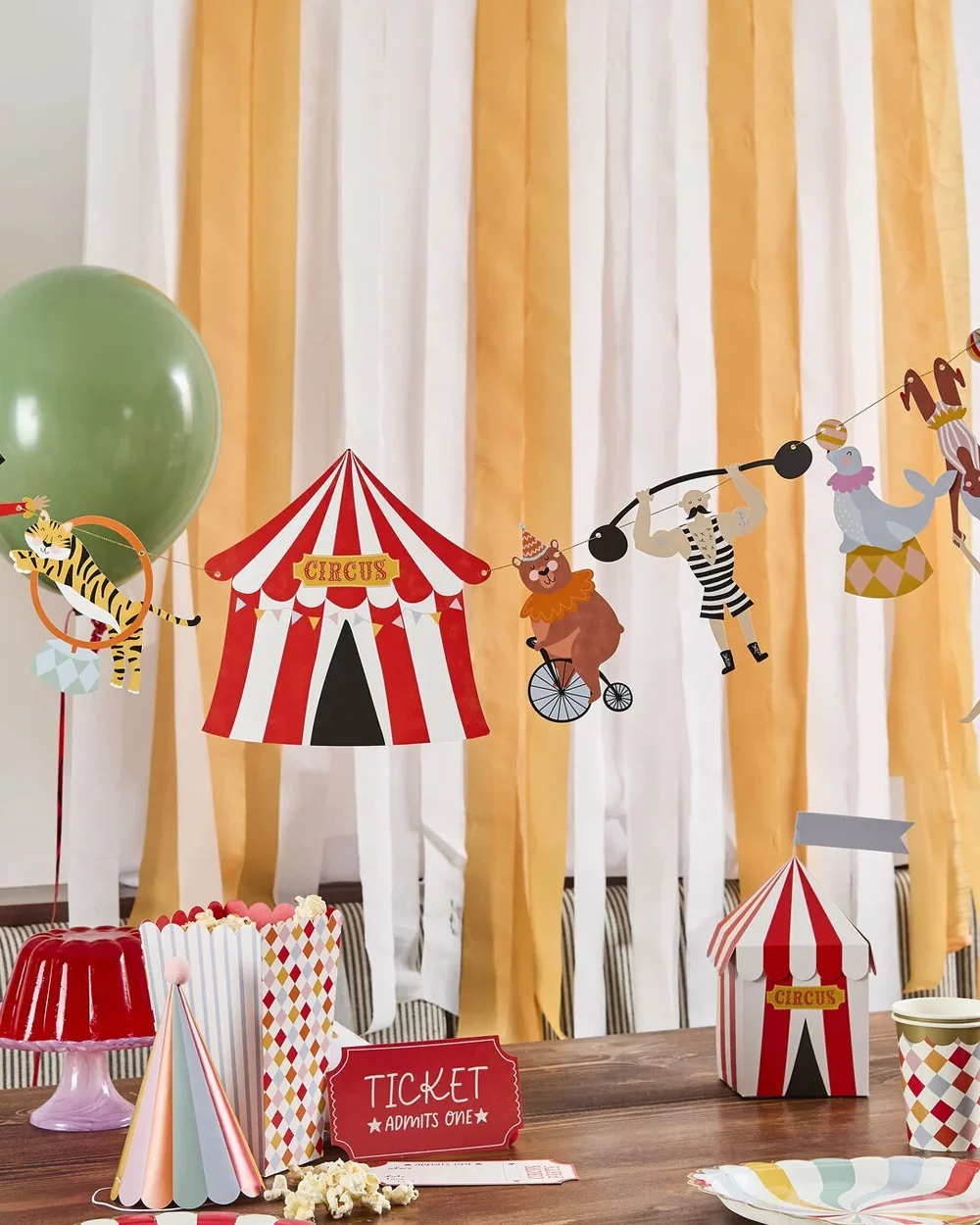 A circus-themed party table display featuring red-and-white striped circus decorations, including a circus tent centrepiece, a circus tent favour box, popcorn containers, patterned paper cups and a ‘Ticket Admits One’ sign. Above the table hangs a garland with illustrated circus performers and animals, including a tiger, bear, seal and strongman, set against a backdrop of gold and white streamers.