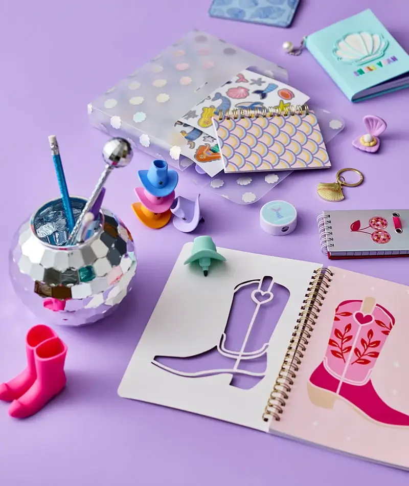 Western-themed stationery set with colourful notebooks, mini cowboy boot accessories, pens, stickers, and novelty desk items arranged on a pastel purple background. Western-themed stationery set with colourful notebooks, mini cowboy boot accessories, pens, stickers, and novelty desk items arranged on a pastel purple background.