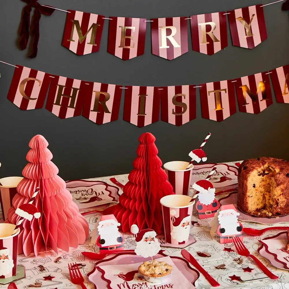 Festive table decorated with red and pink paper Christmas trees, Santa-themed cups and plates, and a large cake, with a red and gold Merry Christmas banner hanging above.