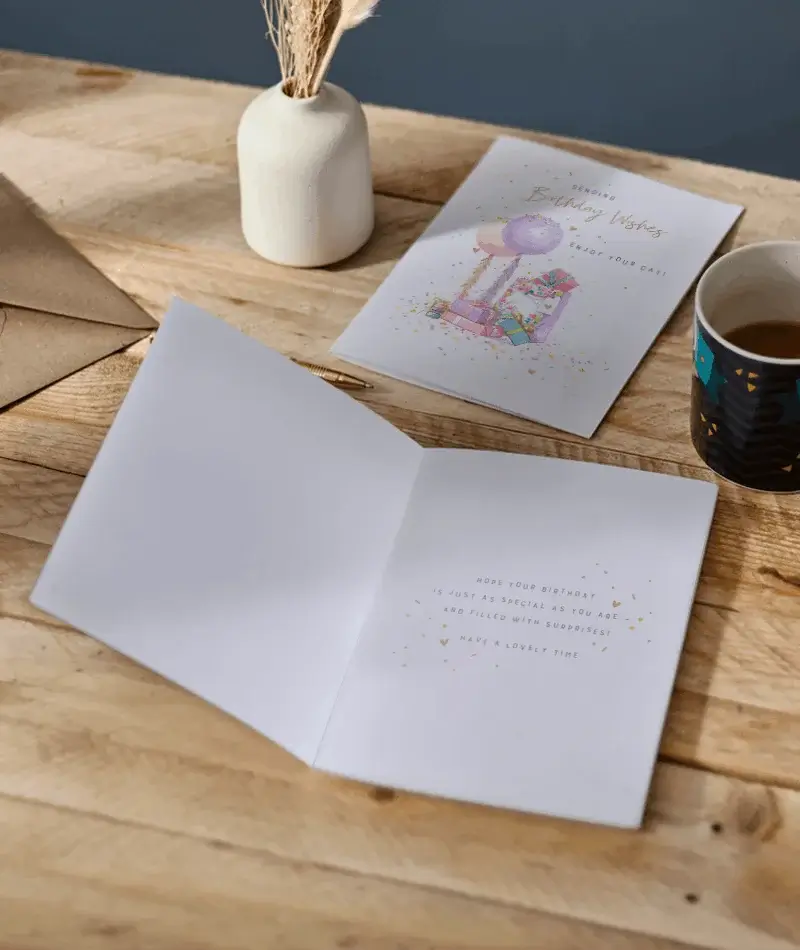 Open birthday greeting card with a printed birthday verse displayed on a wooden table beside the matching card front, envelopes, and a cup of tea. Open birthday greeting card with a printed birthday verse displayed on a wooden table beside the matching card front, envelopes, and a cup of tea.