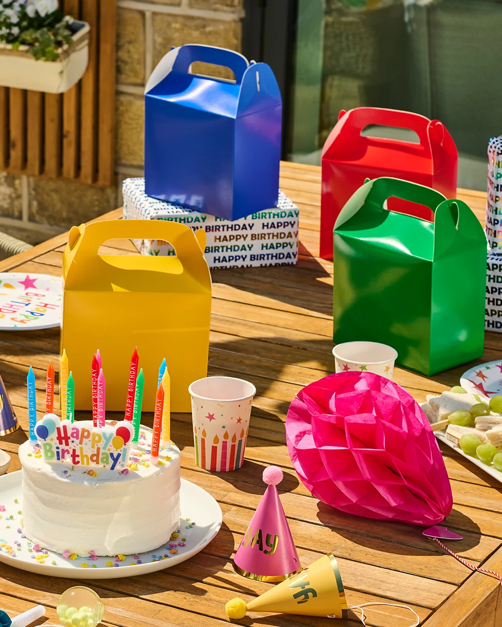 birthday party table display featuring colourful party favour boxes in yellow, blue, red and green, arranged around a birthday cake topped with multicoloured candles and a ‘Happy Birthday’ cake topper. The table is decorated with party hats, cups, snacks and honeycomb paper decorations, creating a bright and colourful celebration setup. birthday party table display featuring colourful party favour boxes in yellow, blue, red and green, arranged around a birthday cake topped with multicoloured candles and a ‘Happy Birthday’ cake topper. The table is decorated with party hats, cups, snacks and honeycomb paper decorations, creating a bright and colourful celebration setup.