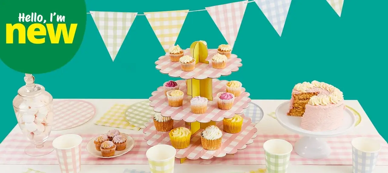 The image shows a colorful and festive dessert table setup. There is a three-tiered pink and white checkered cupcake stand in the center, holding various cupcakes with different colored frostings. Surrounding the stand are pastel-colored cups, plates, and napkins with checkered patterns. On the right side of the table, there is a partially sliced cake with pink frosting on a white cake stand. In the background, there are pastel-colored triangular bunting flags hanging against a teal wall. On the left side of the image, there is text in a green circle that reads 