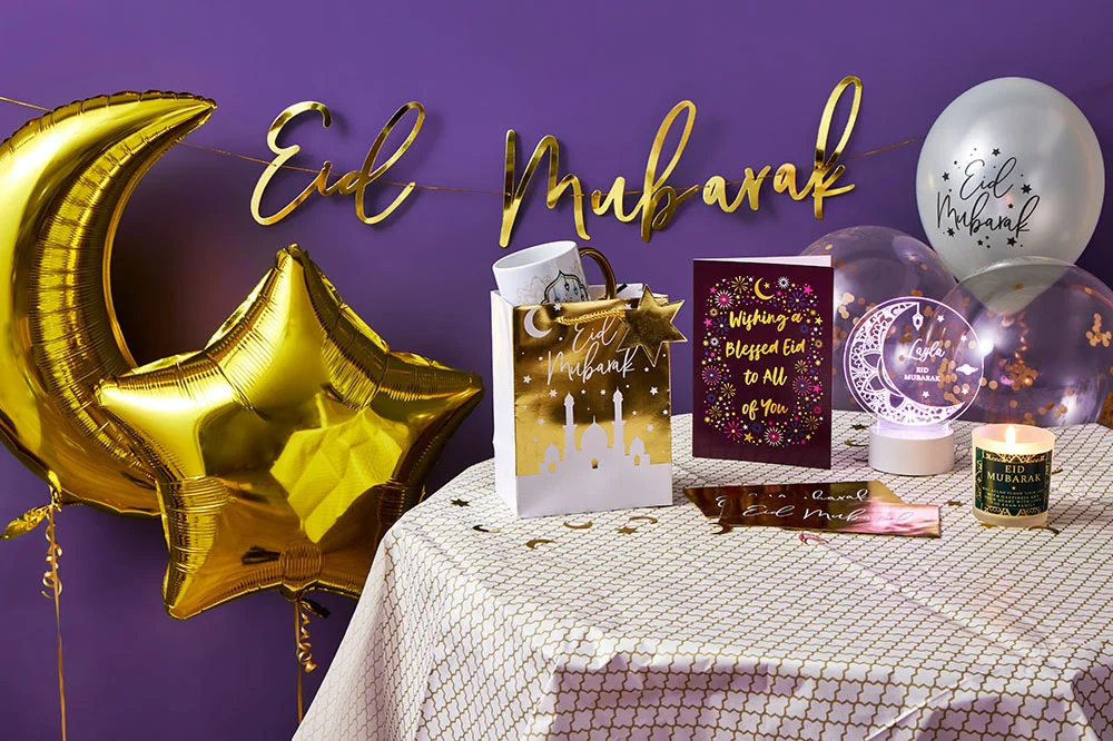 A decorated table with Eid‑themed gifts and decorations, including gold crescent‑moon and star balloons, a gold ‘Eid Mubarak’ banner, a gift bag, greeting cards, an acrylic light ornament, a mug, and a lit candle, set against a purple backdrop.