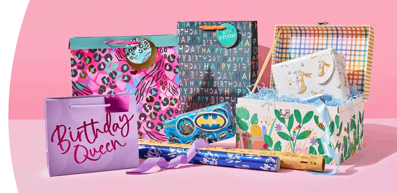Colourful selection of birthday gift bags, boxes, and wrap displayed against a pink background.