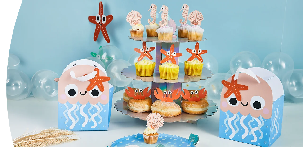 An under‑the‑sea themed party display on a light blue background, featuring a three‑tier cupcake stand with decorated cupcakes, crab‑topped donuts, jellyfish‑shaped party boxes, starfish decorations, shells, and transparent balloons arranged around the table.