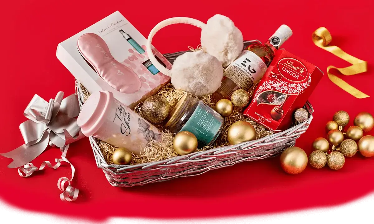 Gift hamper on a red background containing earmuffs, a pink water bottle, a candle, Lindt chocolates, a small bottle of gin, and festive gold ornaments. Gift hamper on a red background containing earmuffs, a pink water bottle, a candle, Lindt chocolates, a small bottle of gin, and festive gold ornaments.