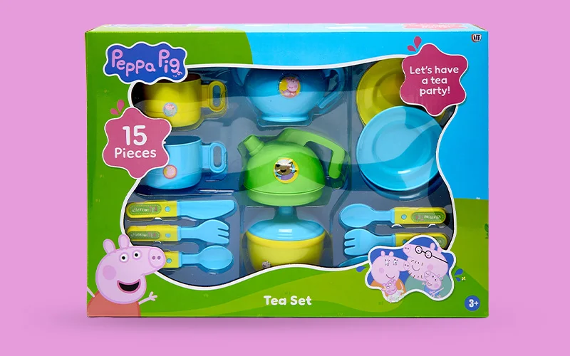 A boxed Peppa Pig children’s tea set containing 15 plastic pieces, including cups, plates, cutlery and a teapot in blue, green and yellow, shown on a pink background.