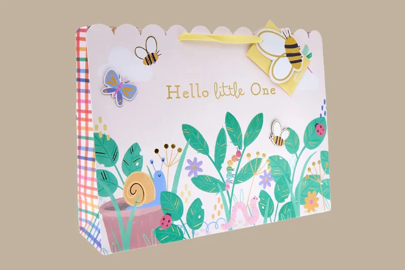 A baby gift bag decorated with colourful garden-themed illustrations including flowers, leaves, bees, butterflies, a snail and ladybirds. The bag features gold lettering reading ‘Hello Little One’ and has yellow ribbon handles.