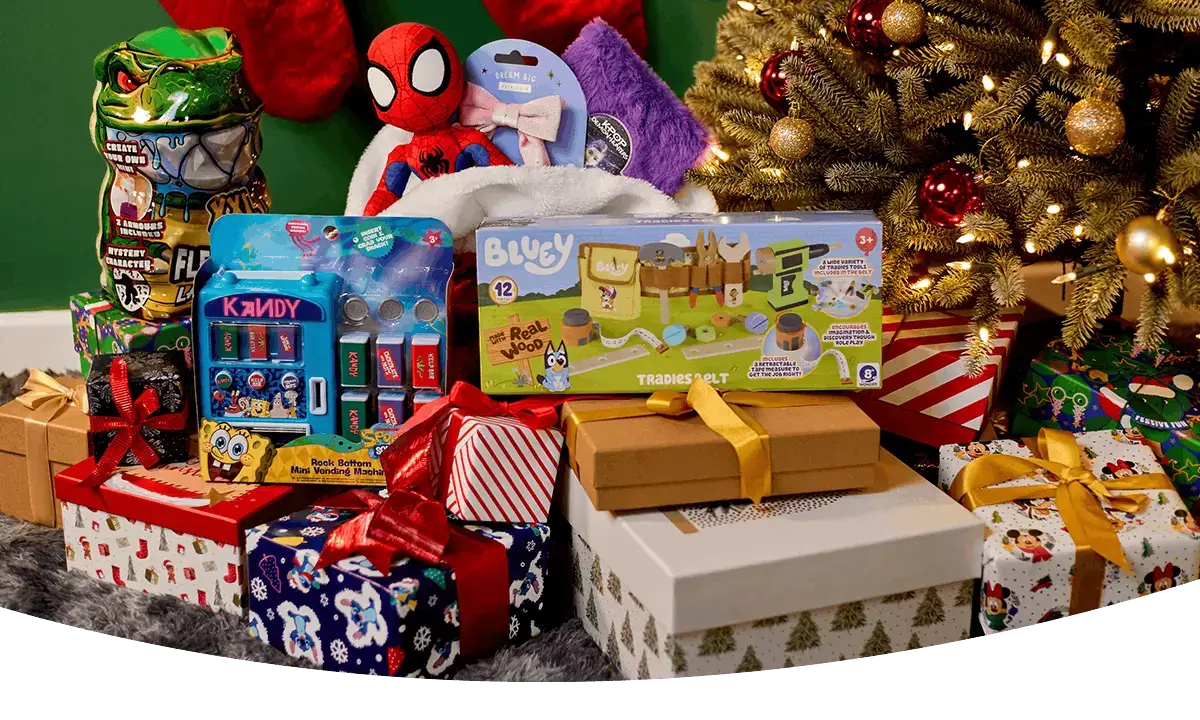 Selection of Christmas gifts and toys, including a Bluey playset, Spider-Man plush, novelty sweets and a miniature vending machine, displayed among wrapped presents beside a decorated Christmas tree.
