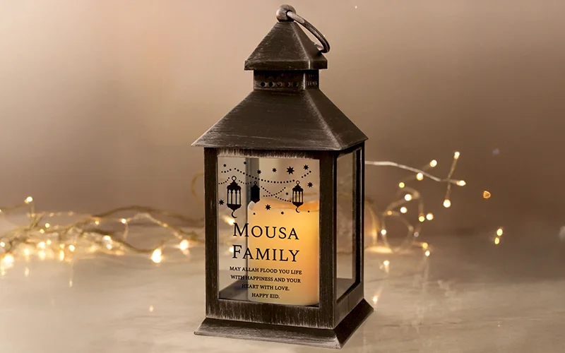 A black metal lantern with a warm glowing candle inside, featuring a decorative panel printed with stars, lanterns, and the text ‘Mousa Family’ along with an Eid blessing.