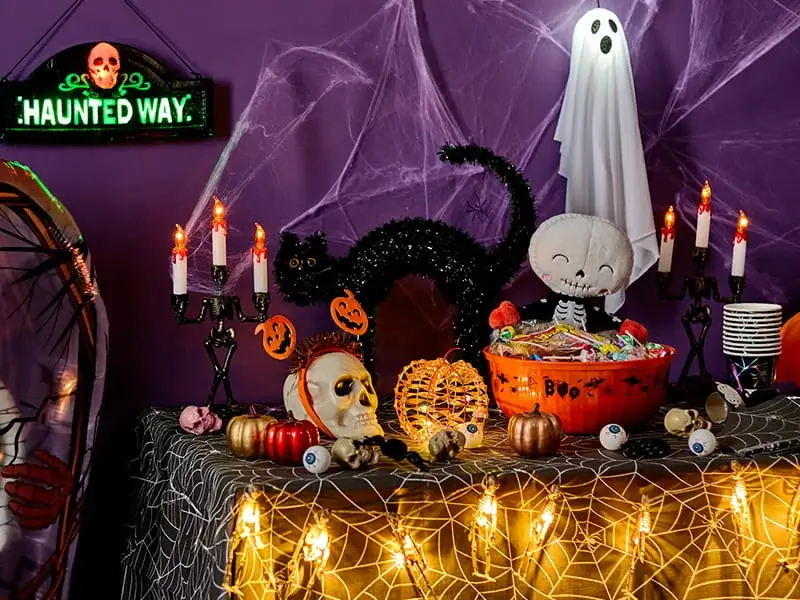 Halloween display featuring a ghost decoration, black cat ornament, trick-or-treat sweets, spiderweb decorations, pumpkins and spooky candle holders against a purple backdrop.