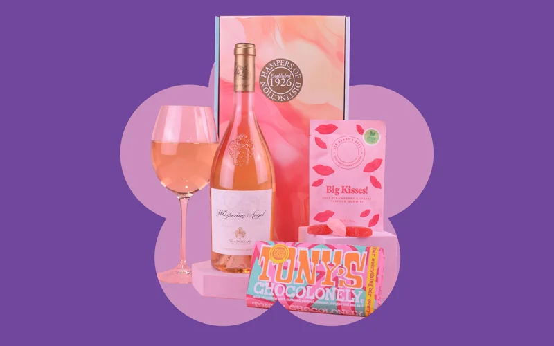 A gift set containing a bottle of rosé wine, a filled wine glass, chocolates, and sweets, arranged in front of a decorative gift box on a purple background with a pink flower shape.