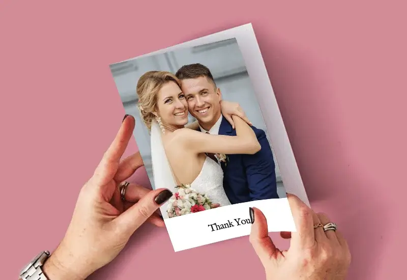 A person holding a thank you card with an illustration of a couple embracing, one in a white dress and the other in a dark suit, on a pink background with Thank You written at the bottom.