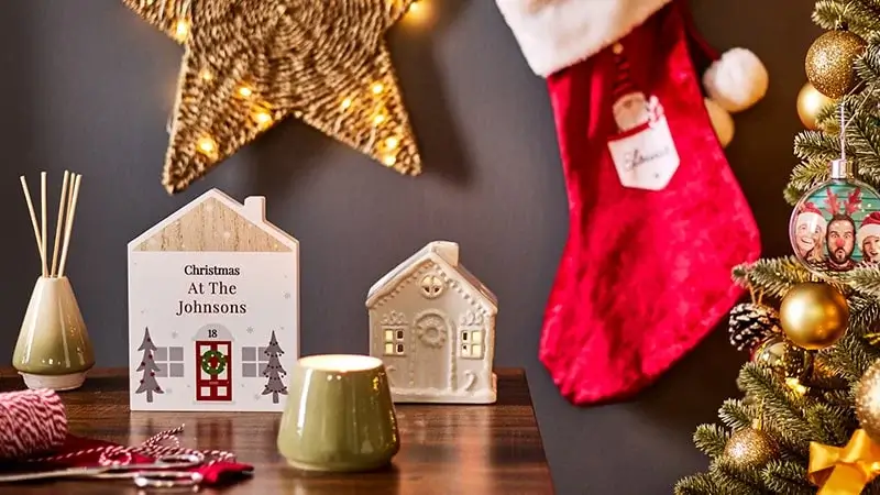 Festive Christmas scene with a decorated tree, gold ornaments, a red stocking, a lit candle, and ceramic house decorations on a wooden table.