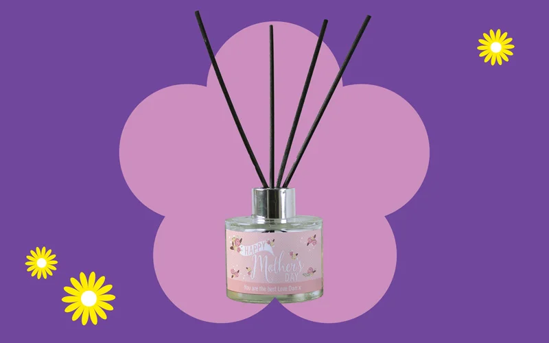 A pink Mother’s Day reed diffuser with black reeds and butterfly decorations, shown against a purple background with a pink flower shape and yellow daisies.