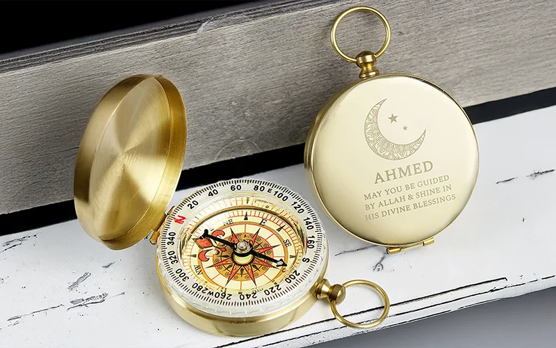 A gold pocket compass with its lid open, showing an ornate compass face, alongside the engraved lid reading ‘Ahmed’ with a crescent moon and star design and a blessing message.