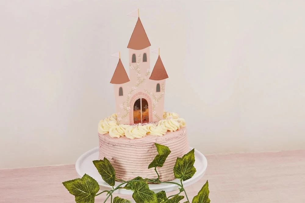 A fairytale castle cake topper in pastel pink with three turrets, floral decorations and miniature flag details, positioned on a pink birthday cake decorated with buttercream swirls and pearl-like sprinkles. A fairytale castle cake topper in pastel pink with three turrets, floral decorations and miniature flag details, positioned on a pink birthday cake decorated with buttercream swirls and pearl-like sprinkles.
