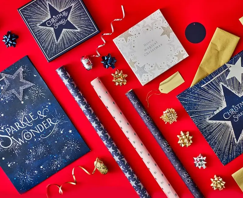 Christmas gift wrapping set displayed on a red background, featuring navy and gold gift bags with star designs and text Sparkle and Wonder and Christmas Sparkle, coordinating wrapping paper rolls in blue and pink patterns, gold envelopes, decorative bows, and curling ribbons.