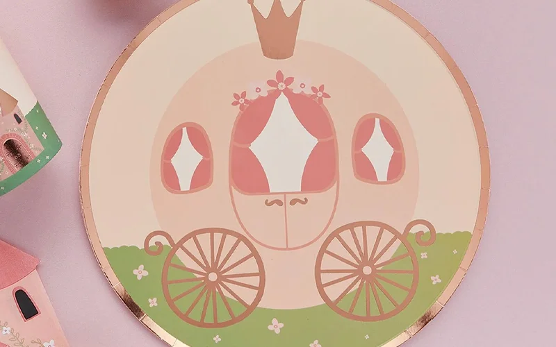 A round princess party plate decorated with an illustrated fairytale carriage, a crown motif, pastel pink and green details, and a gold foil rim, displayed on a pale pink background. A round princess party plate decorated with an illustrated fairytale carriage, a crown motif, pastel pink and green details, and a gold foil rim, displayed on a pale pink background.