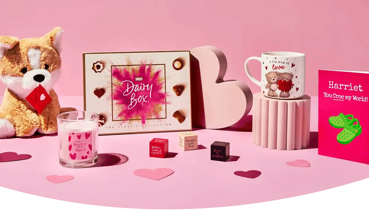 A pink Valentine’s‑themed display featuring a plush dog, a box of chocolates, a heart‑decorated candle, small gift cubes, a cute illustrated mug, and a bright greeting card arranged with scattered paper hearts.
