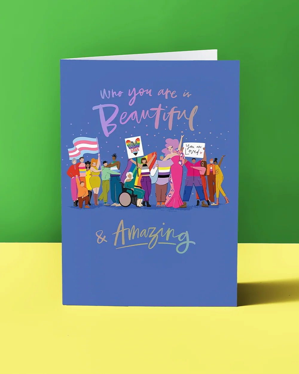 A blue Pride greeting card featuring a group illustration of diverse people holding flags and signs, with text reading ‘Who you are is Beautiful & Amazing,’ displayed against a green and yellow background.