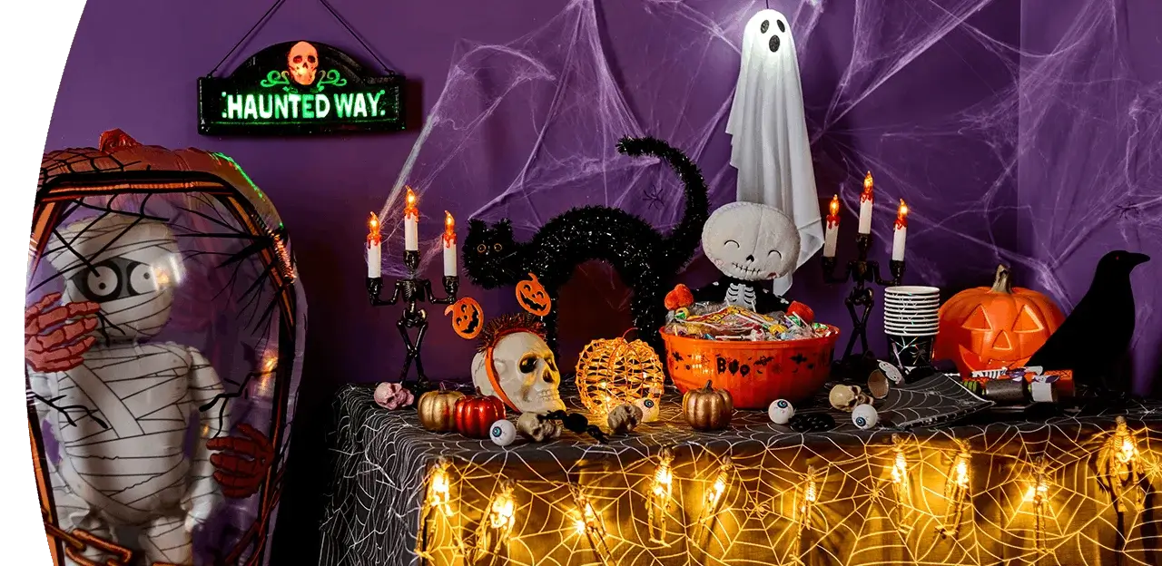 Halloween party table with spooky decorations, trick-or-treat sweets, glowing candles, pumpkins, cobwebs and a mummy figure against a purple backdrop.