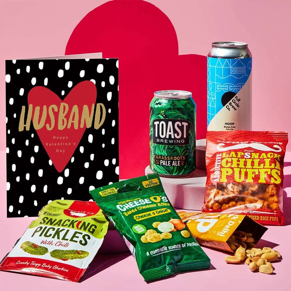 Valentine’s gift set displayed on a pink background with red heart-shaped props. The set includes a brown card with the text I ❤️ your and a rooster illustration, two cans of craft beer, and assorted snacks such as snacking pickles, cheese bites, chili puffs, and nuts in colorful packaging.