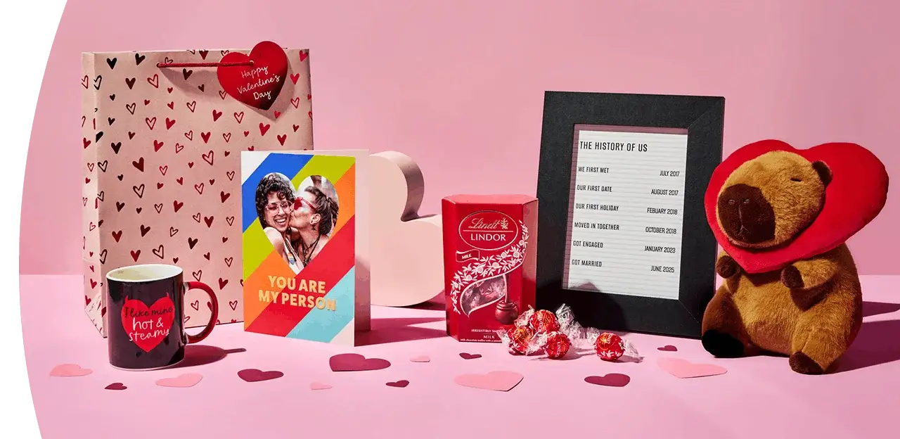 A display of Valentine’s Day gifts including a heart-patterned gift bag, a mug, a colorful greeting card, Lindor chocolates, a framed milestone list, and a plush toy with a red heart behind it, arranged on a pink background with scattered paper hearts.